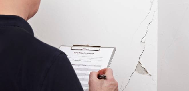 Man with inspection checklist in front of a white wall with a long crack or rip and a piece of plaster missing